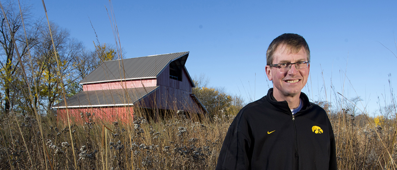 A man stands amidst prairie grasses in front a restored barn